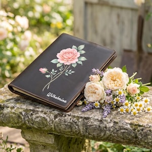 May include: A dark brown leather journal featuring a printed illustration of a pink rose with green leaves and the name Wilhelmina in white script. The journal is positioned next to a bouquet of cream-colored roses and small white daisies.