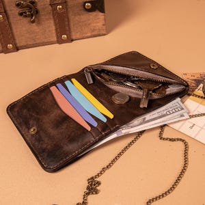 May include: A dark brown leather wallet with a metal chain, featuring a zippered coin compartment, multiple card slots containing pink, blue, and yellow inserts, and a section holding United States one hundred dollar bills and metal coins.