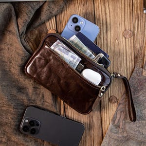 Könnte beinhalten: Eine braune Ledergeldbörse mit Reißverschluss, geöffnet, um Bargeld, einen USB-Stick und weiße Ohrhörer zu zeigen. Ein blaues Smartphone und ein schwarzes Smartphone sind ebenfalls sichtbar. Die Geldbörse liegt auf einer Holzoberfläche.