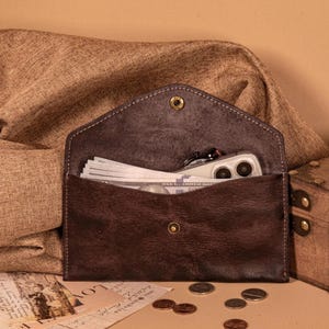 Peut inclure: Un portefeuille en cuir marron foncé de style enveloppe, ouvert pour révéler une liasse de billets de banque américains et un smartphone. Le portefeuille a une fermeture à bouton-pression et une bordure cousue. Quelques pièces et une carte postale vintage sont éparpillées.
