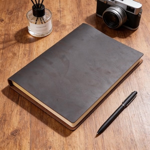 May include: A dark brown leather-bound journal with a pen, a camera, and a glass diffuser bottle with black reeds. The journal is open, revealing cream-colored pages. The pen is black and silver. The items are on a wooden surface.