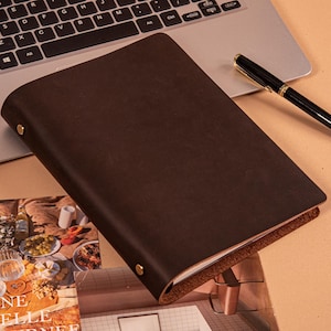 May include: A dark brown leather notebook with a smooth matte finish, featuring two gold-toned metal rivets on the spine edge. The notebook is positioned diagonally on a surface next to a black ballpoint pen and a laptop keyboard.