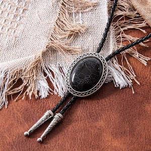 May include: A black bolo tie with a silver-toned oval pendant featuring a cracked stone design. The tie has a braided black cord and decorative silver tips. The bolo tie rests on a brown leather surface.