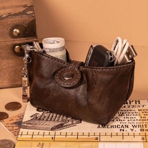 May include: A dark brown leather pouch with a snap button closure, containing a rolled banknote, a black car key fob, and a white USB cable. A silver keychain with a rectangular tag hangs from the side, positioned above a ruler.