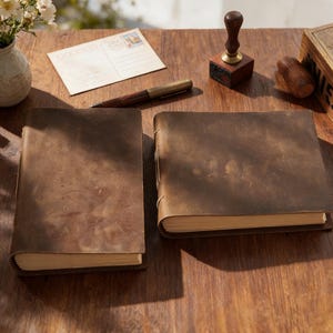 May include: Two brown leather-bound journals with aged paper pages sit on a wooden surface. A postcard, pen, and ink stamp are also on the table. The journals have a rustic, vintage aesthetic.