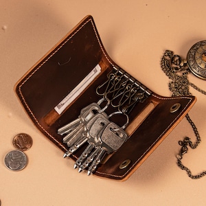 May include: A dark brown leather key case with a snap button closure, featuring six metal key hooks and a small interior slot for folded currency. The case is open, displaying several silver-toned keys attached to the hooks, with two coins nearby.