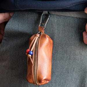 May include: A brown leather key holder with a zipper closure and a metal carabiner clip. The key holder has a small tassel with red and blue beads. The key holder is attached to a gray belt loop.
