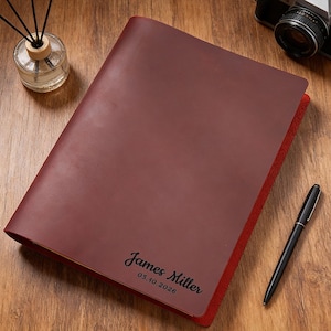 May include: A burgundy leather journal with the name "James Miller" and the date "05.10.2026" printed on the cover. The journal has a red interior and is lying on a wooden surface. A black pen and a camera are also on the surface.