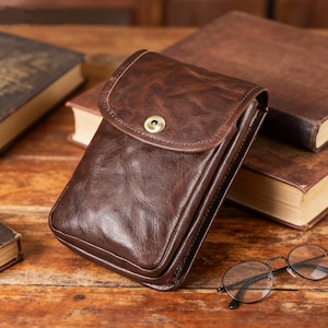 Puede incluir: Una funda de teléfono de cuero marrón con cierre de solapa y un broche de latón. La funda rectangular tiene una superficie texturizada. La funda está sobre una superficie de madera con libros y gafas en el fondo.