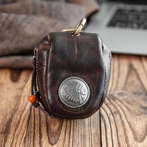 May include: A dark brown leather key pouch with a metal Native American head emblem. The pouch has a zipper closure, a gold-colored metal clasp, and a decorative string with an orange bead. The pouch is on a wooden surface.