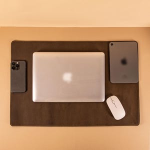 Könnte beinhalten: Eine braune Schreibtischunterlage aus Leder mit einem silbernen Laptop, Tablet, Smartphone und einer weißen Maus. Der Laptop und das Tablet haben das Apple-Logo. Das Smartphone ist schwarz.