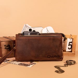 May include: A brown leather clutch bag with a wrist strap and two zippered compartments. The front features the embossed name Owen. The bag is filled with a smartphone, a white computer mouse, and a small black calculator.