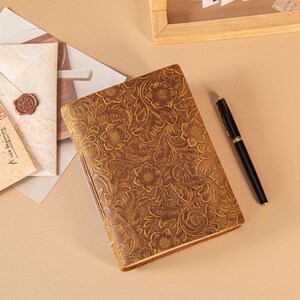 Könnte beinhalten: Ein braunes Lederjournal mit geprägtem Blumenmuster. Ein schwarzer Stift mit goldenen Akzenten liegt neben dem Journal. Das Journal ist geschlossen und hat einen strukturierten Einband. Der Hintergrund ist in einem neutralen Beigeton gehalten.