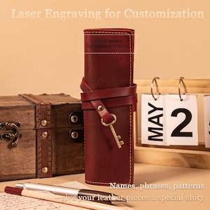 May include: A burgundy leather pen case with a strap and key charm. The case is wrapped with a leather strap and secured with a brass-colored key. Text on the image reads "Laser Engraving for Customization" and "Names, phrases, patterns give your leather pieces a special story."