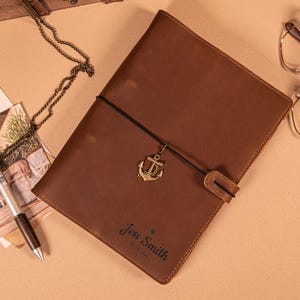 May include: A brown leather journal with an anchor charm and a black elastic closure. The journal is personalized with the name "Jess Smith" and the date "05.10.2026" embossed on the cover. A pen and necklace are visible.
