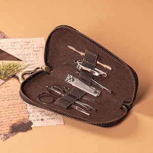 May include: A brown leatherette travel grooming kit with a black zipper, containing various stainless steel manicure tools. The kit is open, revealing the tools secured by brown straps. The background is a neutral tan color.