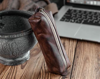 May include: A brown leather pencil case with a silver zipper. The case is cylindrical and sits on a wooden surface. A laptop and a decorative stone bowl are in the background.