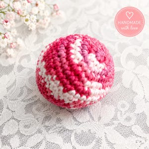 May include: A handmade, crocheted ball in shades of pink and white. The ball has a textured appearance and is set against a white lace background. A pink circular sticker with a heart and the words "handmade with love" is visible.