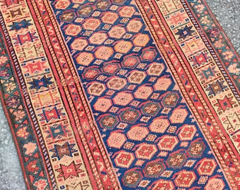 May include: A detailed, rectangular rug with a central blue field featuring a repeating pattern of pink and brown hexagons. The rug has red and cream borders with geometric designs, creating a vibrant, traditional aesthetic.