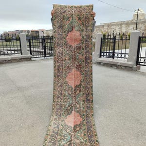 May include: A rectangular, vintage-style rug with a central design in shades of pink and green. The rug has a detailed pattern with a border and is held up against a neutral background.
