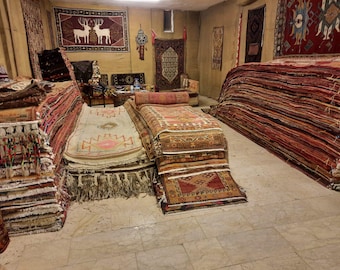 Op de afbeelding: Een grote collectie geweven vloerkleden gestapeld in een kamer, met diverse geometrische patronen, franjes en warme kleurenpaletten waaronder rood, beige en bruin. Verschillende kleden zijn aan de muren opgehangen, waaronder een met herten.