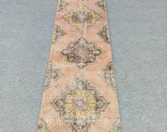Puede incluir: Una alfombra larga y rectangular con una apariencia descolorida y vintage. La alfombra presenta un fondo melocotón suave con patrones de medallones florales repetidos en tonos de marrón, amarillo y verde. La alfombra probablemente esté hecha de lana o un material similar.