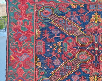 May include: A detailed close-up of a Persian rug with a rich color palette. The rug features a deep blue central field with intricate geometric patterns in red, green, and yellow. A red border frames the design, adding to the rug's traditional aesthetic.