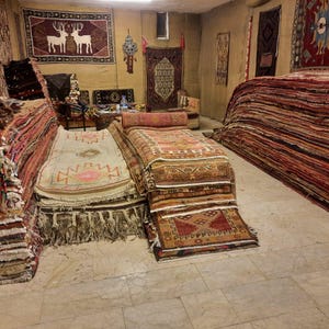 May include: A large collection of woven area rugs stacked in a room, featuring various geometric patterns, fringe edges, and warm color palettes including red, beige, and brown. Several rugs are displayed on the walls, including one with deer.