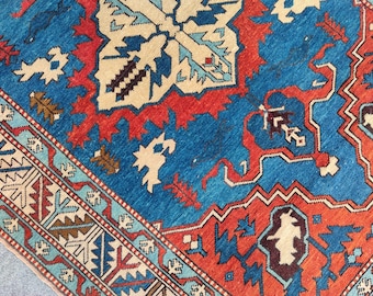 May include: Close-up of a vibrant, patterned rug with a central design in cream, surrounded by a rich blue field. The rug features intricate red and blue geometric borders and accents, creating a visually striking textile.