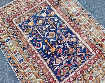 Op de afbeelding: Een antiek tapijt met een centraal blauw veld met een ingewikkeld patroon van geometrische vormen en gestileerde figuren in tinten oranje, geel en wit. Het tapijt wordt omlijst door een rand van rode, bruine en crèmekleurige banden met herhaalde bloemen- en geometrische ontwerpen.