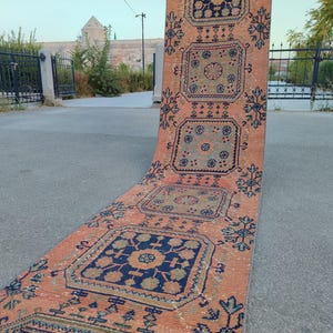 May include: An orange and navy blue runner rug with a geometric pattern. The rug features repeating square and floral designs. The runner is displayed outdoors on a paved surface.