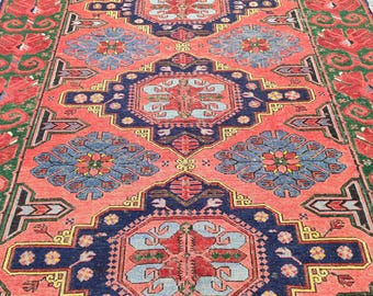 May include: A long, rectangular rug with an intricate pattern. The rug features a coral-colored field with navy blue, light blue, green, and yellow geometric designs. The border is green with red floral motifs.