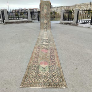 May include: An antique, long, rectangular rug with a detailed pattern. The rug features a central design with floral and geometric motifs in shades of brown, green, pink, and cream. The rug is displayed outdoors on a concrete surface.