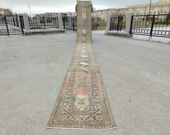 May include: An antique, long, rectangular rug with a detailed pattern. The rug features a central design with floral and geometric motifs in shades of brown, green, pink, and cream. The rug is displayed outdoors on a concrete surface.