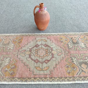 May include: A rectangular woven rug featuring a central geometric medallion in shades of dusty pink, muted orange, and grey, surrounded by a patterned border. Above the rug sits a terracotta clay jug with a single handle and a small tassel.