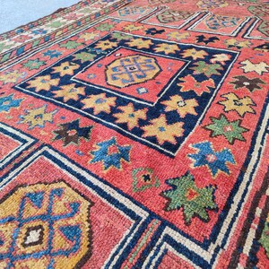 Può includere: Vista dettagliata di un tappeto vintage con un complesso design geometrico. Il tappeto presenta uno sfondo rosso con accenti blu navy, gialli, verdi e marroni. Il motivo include forme geometriche e a stella ripetute.