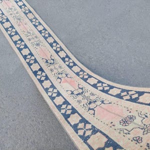 May include: A long, narrow rug with a cream-colored background, featuring a repeating pattern of blue and pink geometric shapes and floral motifs. The rug's design includes a central pink stripe and blue borders.