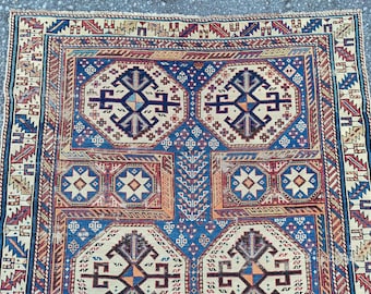Op de afbeelding: Een gedetailleerd vloerkleed met een complex patroon van geometrische vormen in blauw, beige en rood. Het vloerkleed heeft een rand met herhaalde geometrische ontwerpen. Het ontwerp is symmetrisch en gedetailleerd.