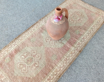 May include: A rectangular runner rug featuring a faded pink and beige geometric pattern with a fringed edge, topped with a terracotta clay pitcher that has a single handle and a decorative white and pink tassel tied around its neck.