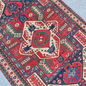Op de afbeelding: Een rechthoekig tapijt met een gedetailleerd geometrisch patroon. Het centrale ontwerp is rood, crème en blauw, omgeven door randen met herhalende motieven. Het ontwerp is symmetrisch en ingewikkeld, wat traditioneel vakmanschap laat zien. Afmetingen in centimeters.