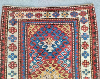 Op de afbeelding: Een rechthoekig geweven tapijt met een centraal rood veld met blauwe en gele geometrische patronen. De rand bestaat uit een crèmekleurige band versierd met herhalende blauwe en rode bloemmotieven, omlijst door donkerbruine randen.