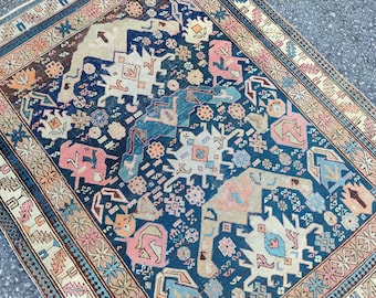 Op de afbeelding: Een antiek tapijt met een gedetailleerd, geometrisch patroon. Het tapijt heeft een diepblauw middenveld met herhalende vormen in roze, beige en blauwgroen. Een patroonrand in complementaire kleuren omlijst het ontwerp. Het ontwerp van het tapijt is ingewikkeld en visueel aantrekkelijk.