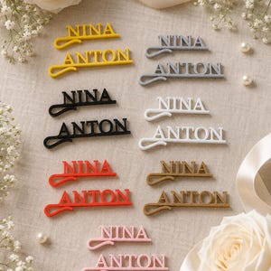 Könnte beinhalten: Eine Sammlung von Haarspangen in verschiedenen Farben, darunter Gelb, Grau, Schwarz, Weiß, Rot, Gold und Rosa. Jede Spange zeigt den Namen NINA oder ANTON in einer Schreibschrift mit einem integrierten Haarnadel-Design an der Basis.