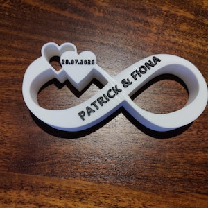 Puede incluir: Un símbolo de infinito blanco con los nombres "PATRICK & FIONA" en texto negro. Un corazón con la fecha "28.07.2020" está integrado en el diseño. El objeto está hecho de un material sólido y descansa sobre una superficie de madera.