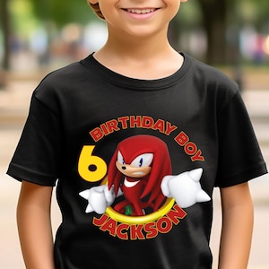 May include: A black short-sleeved t-shirt featuring a graphic of the red character Knuckles from Sonic the Hedgehog, a yellow number 6, and the text "BIRTHDAY BOY" and "JACKSON" printed in a circular arrangement around the character.