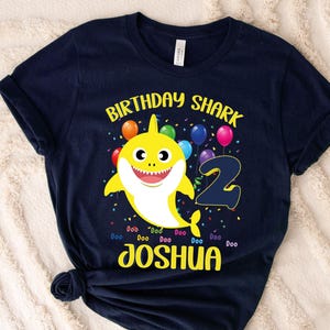 May include: Navy blue t-shirt with a yellow shark graphic, colorful balloons, and the text "Birthday Shark 2 Joshua". The shark has a smiling face and is surrounded by confetti.