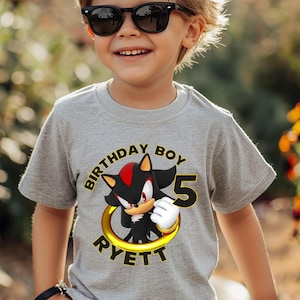 May include: A light gray t-shirt featuring a cartoon character with the text "BIRTHDAY BOY 5 RYETT". The graphic includes a black and red character inside a gold ring. The boy is wearing black sunglasses.