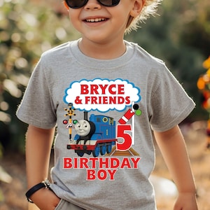 May include: A gray t-shirt featuring a blue train with the text "Bryce & Friends" and "Birthday Boy". The number "5" is prominently displayed. The shirt is worn by a child wearing sunglasses.