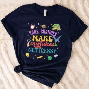 May include: A navy blue t-shirt with the slogan "Take Chances, Make Mistakes, Get Messy" in a retro font. The design includes illustrations of a clock, planet, dinosaur, and other school-related images.
