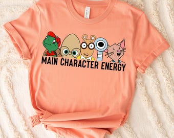 Op de afbeelding: Perzikkleurig T-shirt met stripfiguren en de tekst "MAIN CHARACTER ENERGY" in het zwart. De afbeelding bevat een schildpad, een eivormig personage, Arthur, een duif en een kat. Het shirt heeft korte mouwen en een ronde hals.